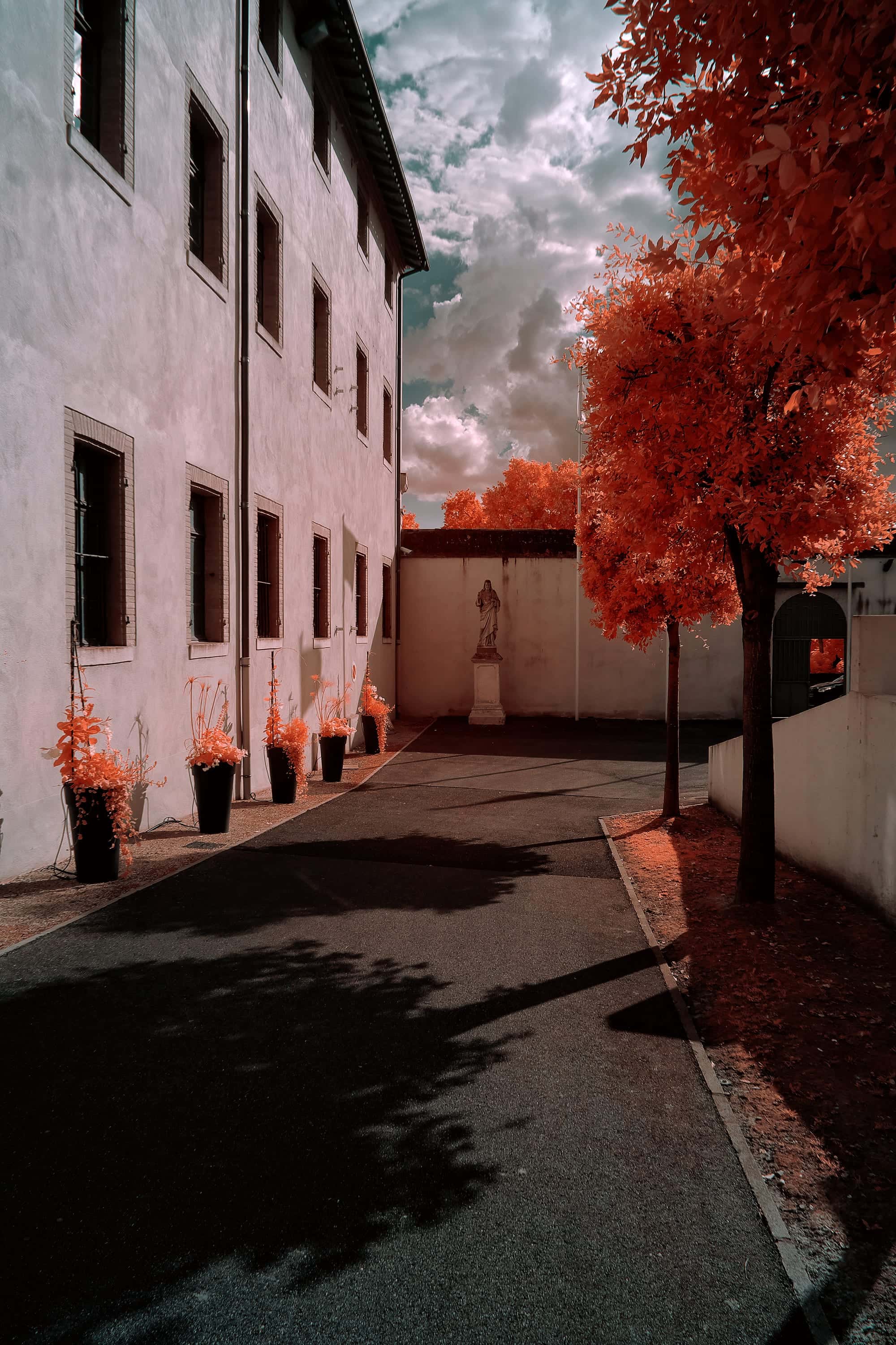 A robed figure on a pedestal at the closed end of a lane between a shuttered building and a garden wall in France, photographed in near-infrared light.