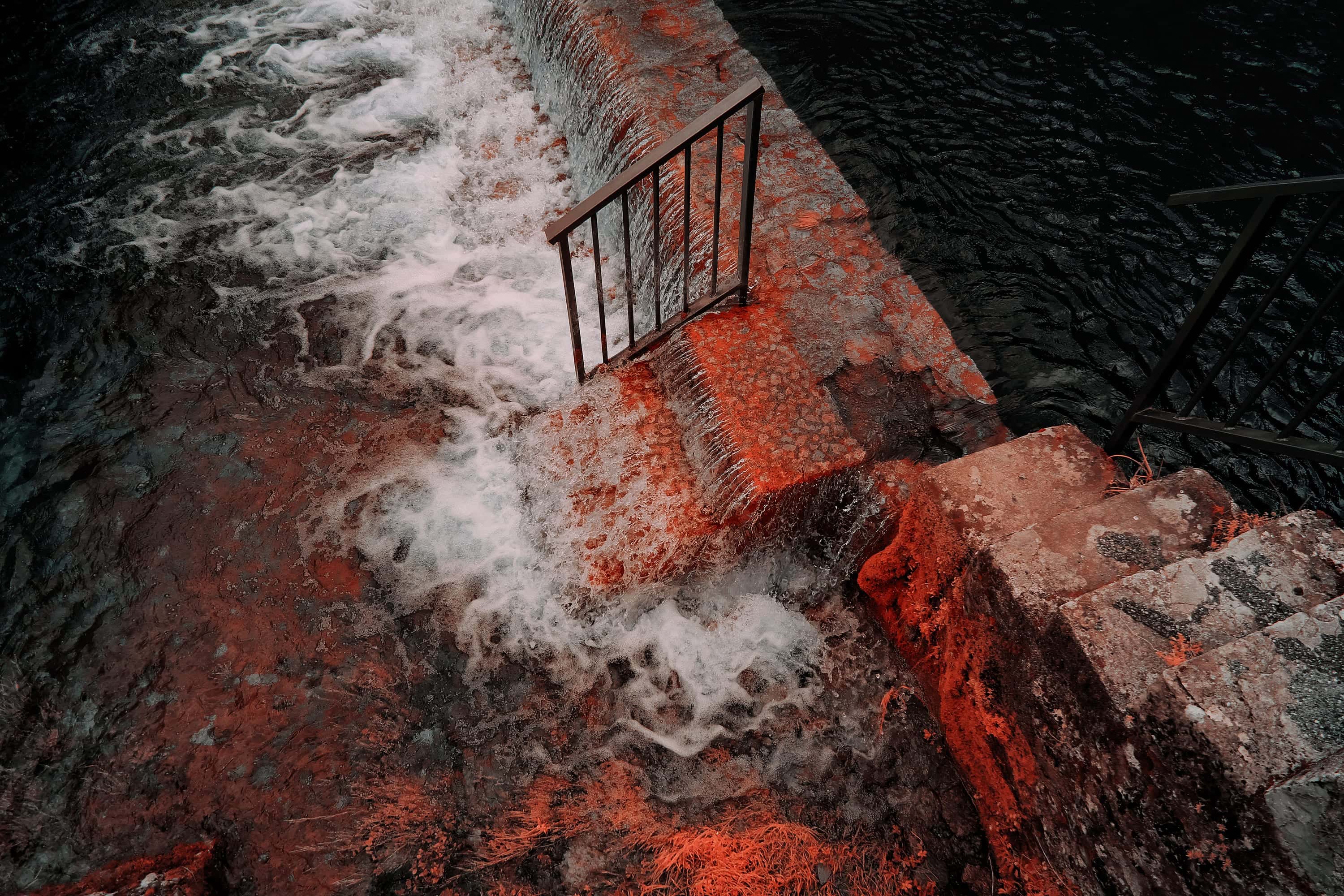 Water spilling over a low stone weir beside a handrail and a flight of steps, photographed in near-infrared light.