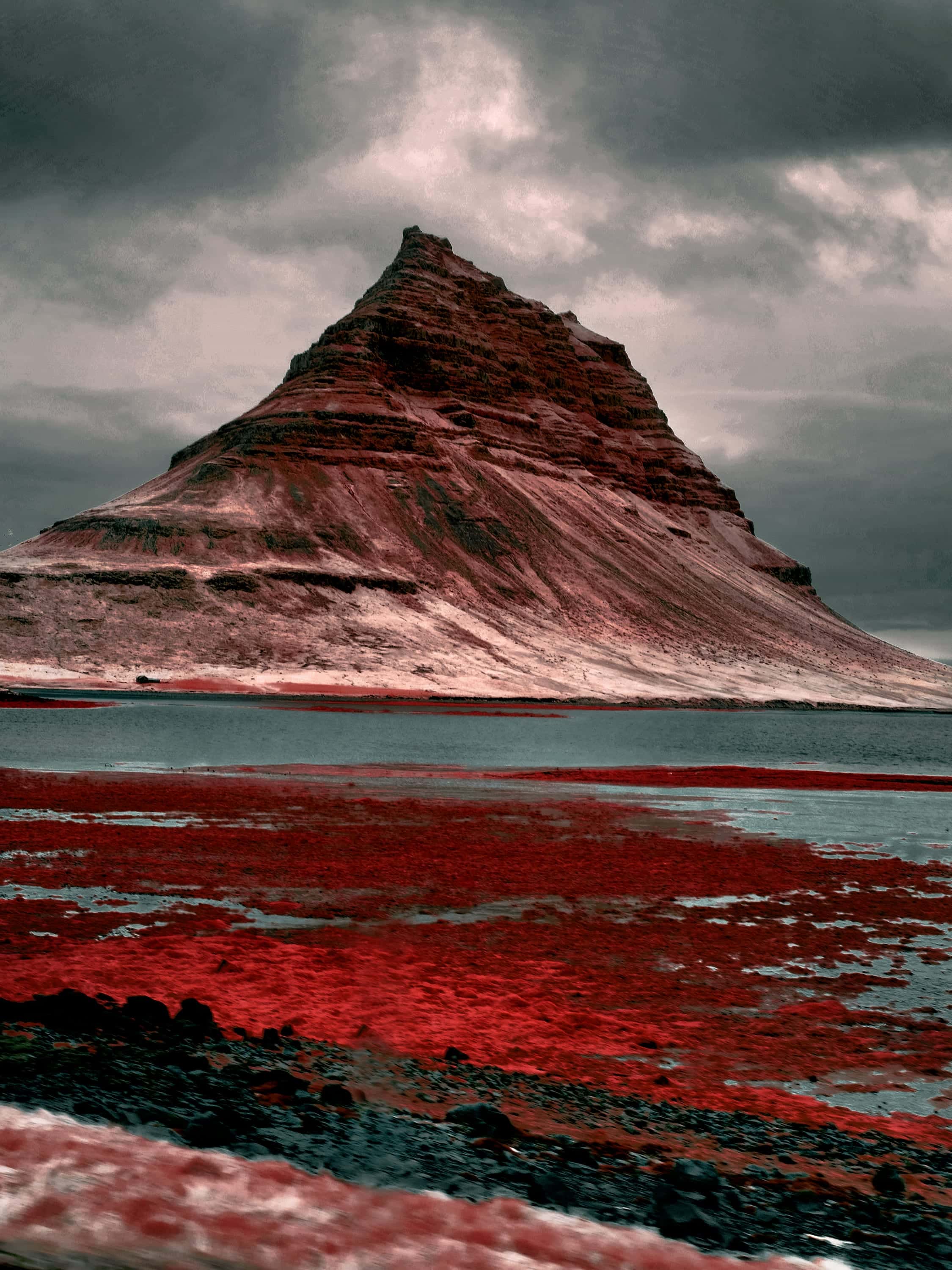 Kirkjufell rising in banded layers under heavy cloud, the tidal flat below it blazing crimson