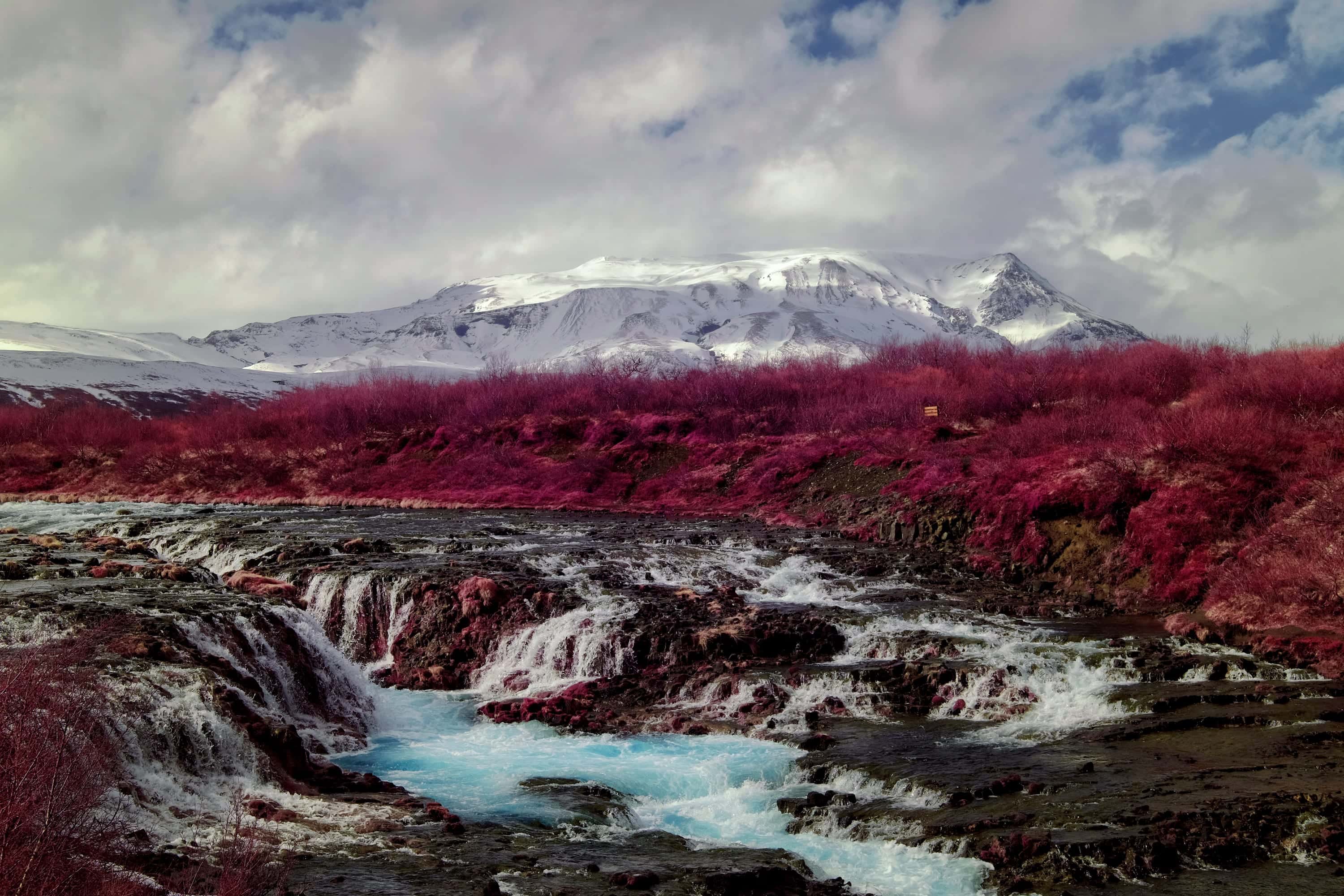 Braided cascades stepping over a broad dark ledge into a fast channel, with a snow-covered massif behind, photographed in near-infrared light.