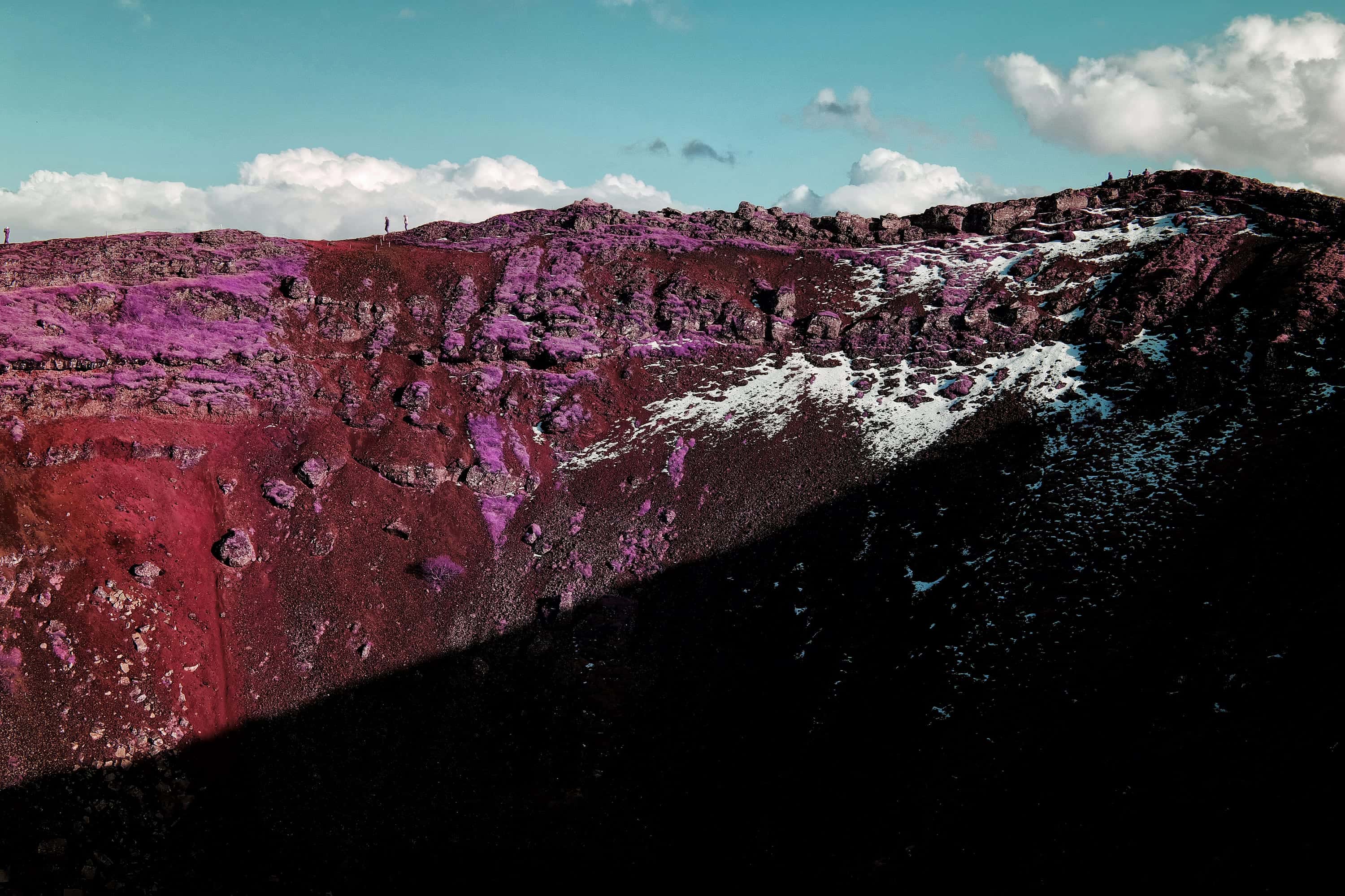The inner wall of a volcanic crater in loose scoria, patched with moss and snow, with people walking the rim above, photographed in near-infrared light.
