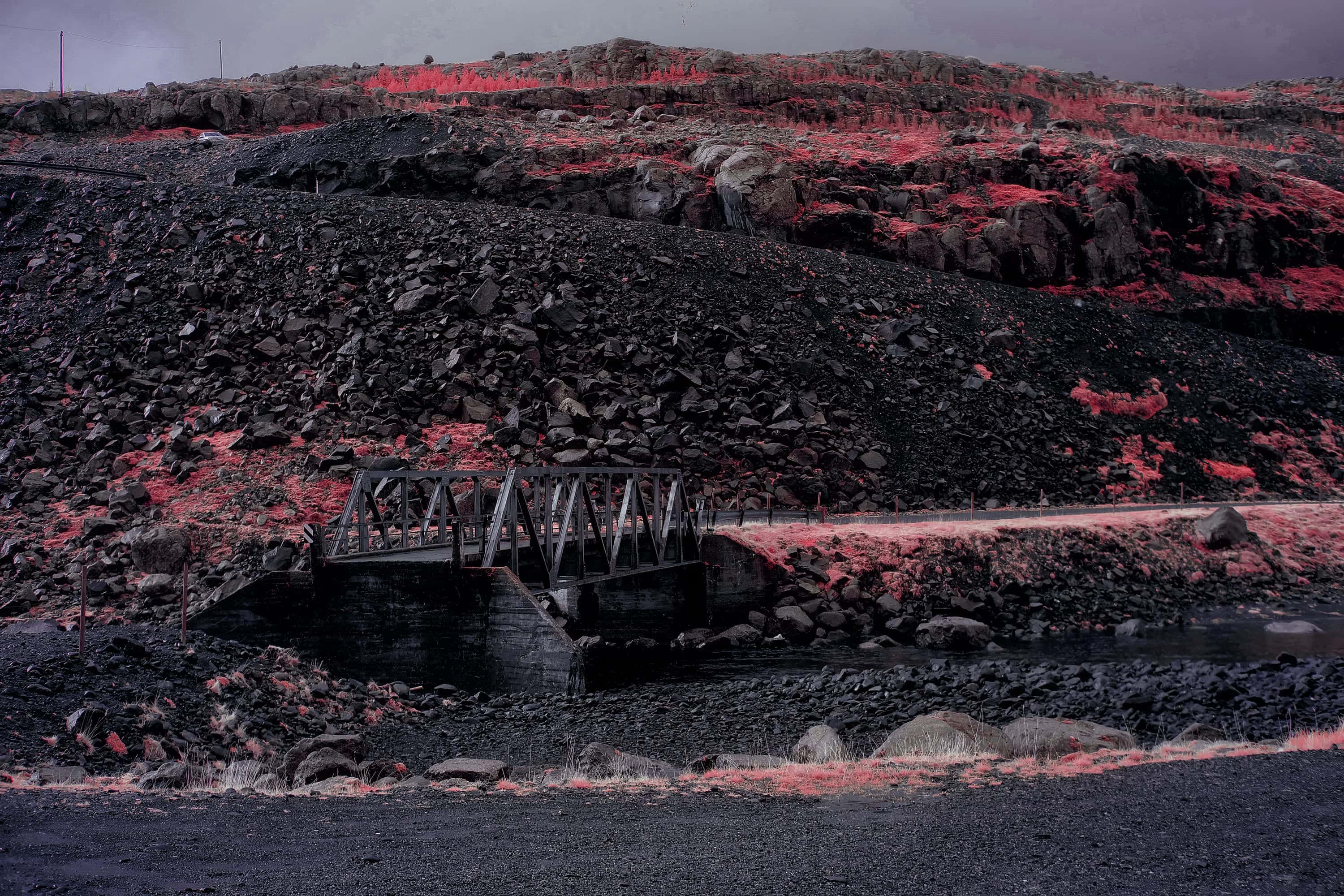 An old single-lane steel truss bridge crossing a river below a slope of dark scree, photographed in near-infrared light.
