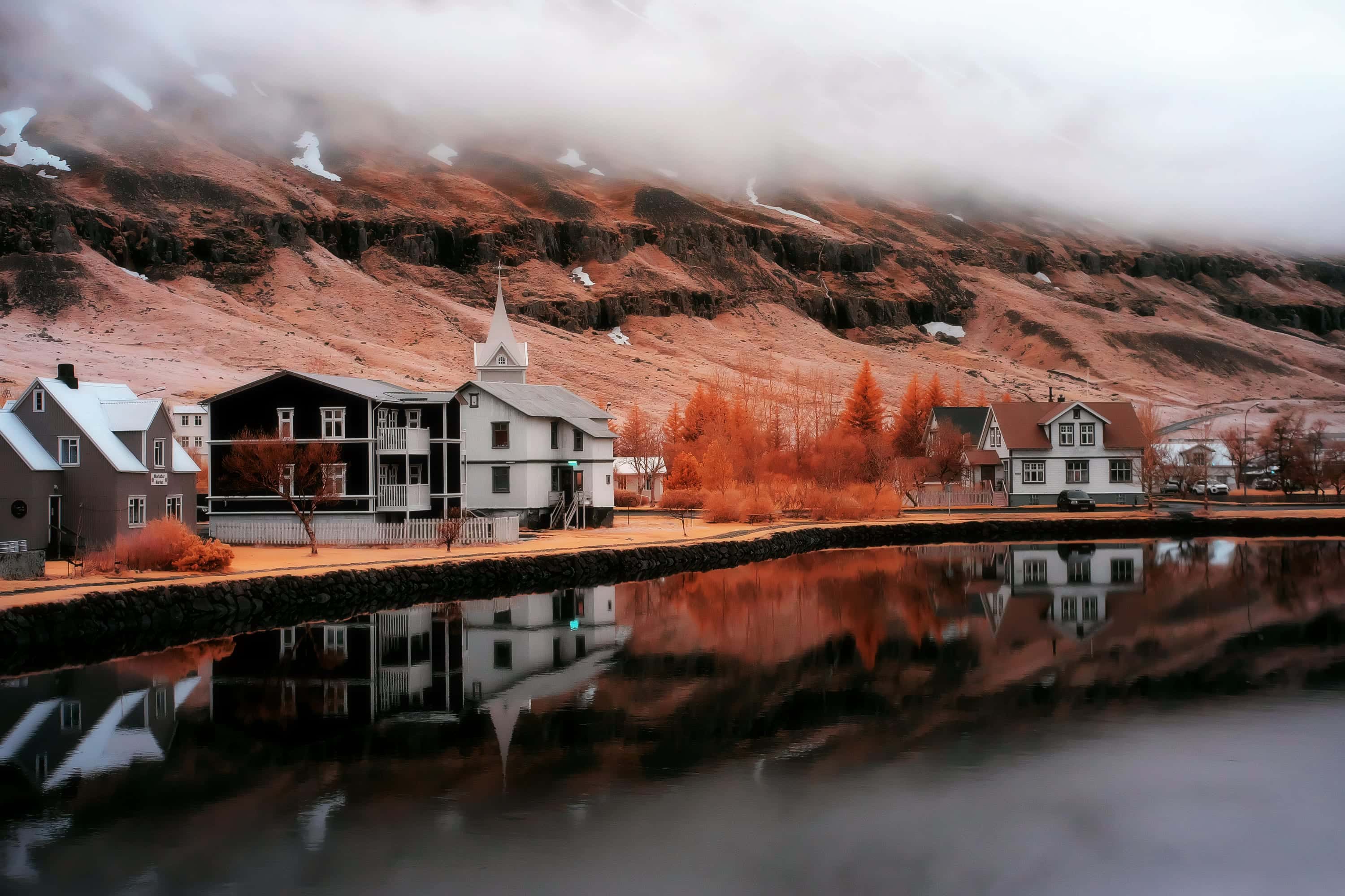 A timber church and painted houses reflected in the still lagoon at the head of an Icelandic fjord, photographed in near-infrared light.