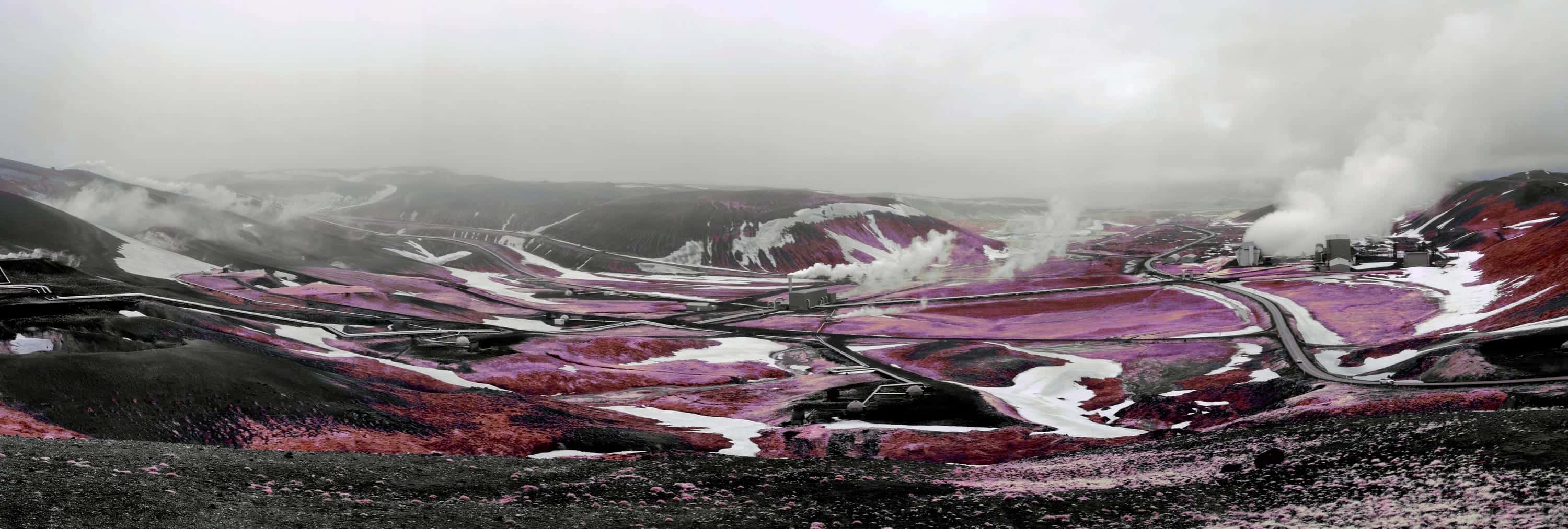 A geothermal power station and its wellhead pipelines spread across a snow-patched basin, with steam rising from several points, photographed in near-infrared light.
